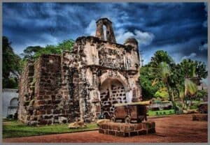 This image shows the historical remnants of A Famosa, a Portuguese fortress located in Malacca, Malaysia. In the foreground, there are two old cannons mounted on carriages, positioned on a brick-laid area. The background features the weathered gatehouse, with its arched entrance and a partial ruin of a watchtower above, exhibiting signs of its age and battles it has witnessed. Tropical vegetation flanks the site, and the sky above is dramatic, with a mix of blue tones and scattered clouds, indicating it might be a HDR (High Dynamic Range) photograph. The overall atmosphere is one of historical significance, blending the bygone era with the present day.