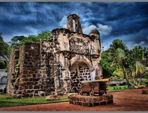 This image shows the historical remnants of A Famosa, a Portuguese fortress located in Malacca, Malaysia. In the foreground, there are two old cannons mounted on carriages, positioned on a brick-laid area. The background features the weathered gatehouse, with its arched entrance and a partial ruin of a watchtower above, exhibiting signs of its age and battles it has witnessed. Tropical vegetation flanks the site, and the sky above is dramatic, with a mix of blue tones and scattered clouds, indicating it might be a HDR (High Dynamic Range) photograph. The overall atmosphere is one of historical significance, blending the bygone era with the present day.