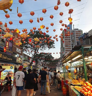 Colorful lanterns and bustling street market scene during the day in an Asian city, featuring food stalls, shoppers, and vibrant decorations. Perfect for highlighting cultural markets, street food, and festive atmosphere.