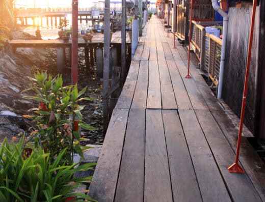 A scenic wooden pathway along a waterfront with red lanterns hanging overhead and lush green plants on the side, creating a serene and traditional Asian ambiance during sunset.