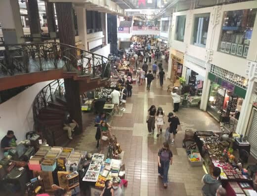 Colorful indoor marketplace with various stalls and shoppers browsing, featuring a staircase on the left side and multiple retail shops promoting arts, crafts, and souvenirs.