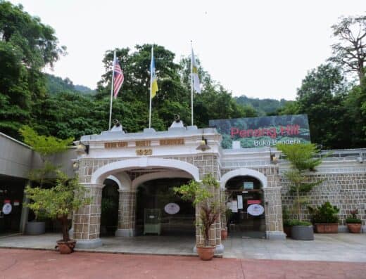 槟城山武吉本德拉历史入口大门建于 1923 年，周围绿树环绕，旗帜飘扬，展现了槟城著名山坡景点的标志性景观。.