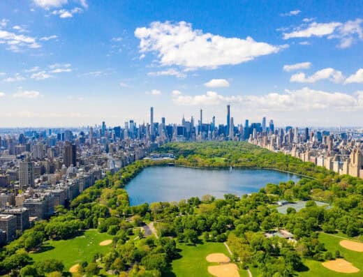 Vibrant aerial view of Central Park in New York City with lush greenery, scenic lake, and iconic skyline skyscrapers under a bright blue sky. Perfect for travel and urban park-related content.