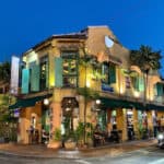 Vibrant yellow and green tropical-style building with outdoor seating, lush palm plants, and colorful lighting at dusk, representing a lively restaurant or cafe in a tropical location.