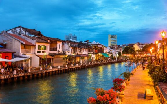 Scenic view of vibrant riverside street in Onalulu with historic buildings, lively cafes, colorful flowers, and glowing street lamps during evening, showcasing charming urban waterfront atmosphere.