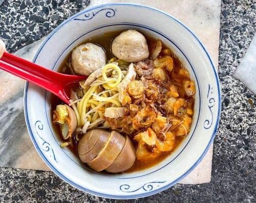 A comforting bowl of Asian-style spicy noodle soup featuring meatballs, noodles, and savory broth, served in a white bowl with a red spoon. Perfect for Asian cuisine lovers.