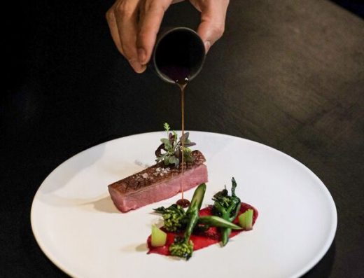 Close-up of a chef pouring rich sauce over a perfectly cooked, medium-rare steak garnished with herbs, served with colorful vegetables on a sleek white plate, emphasizing gourmet cuisine and fine dining experience.