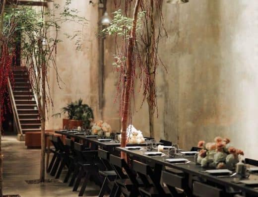 Beautiful industrial restaurant interior featuring a long dining table, modern black chairs, lush plants, hanging lights, and floral centerpieces, ideal for upscale dining experiences.