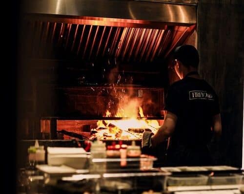 A chef cooking on an open flame grill in a professional kitchen, emphasizing grilling techniques and fire control for delicious dishes.