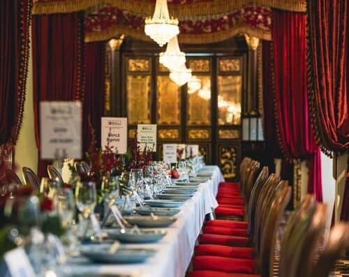豪华宴会厅内布置了精美的瓷器、玻璃器皿和花卉中心装饰品，厅内悬挂着枝形吊灯，并装饰着华丽的帷幔。.