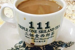 特写镜头：一个装满热茶的白色陶瓷杯，杯身上印有黑色和绿色的"Triple One Sri Land Tea"标志，放置在一块背景模糊的装饰布上。.