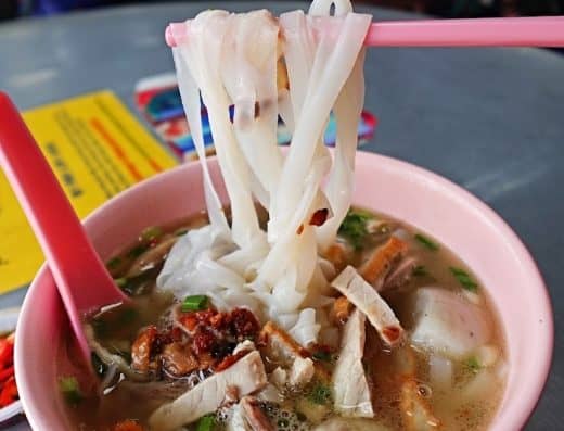 Steaming bowl of traditional Asian chicken noodle soup with fresh herbs, tender meat, and rice noodles in a cozy restaurant setting, perfect for comfort food lovers.