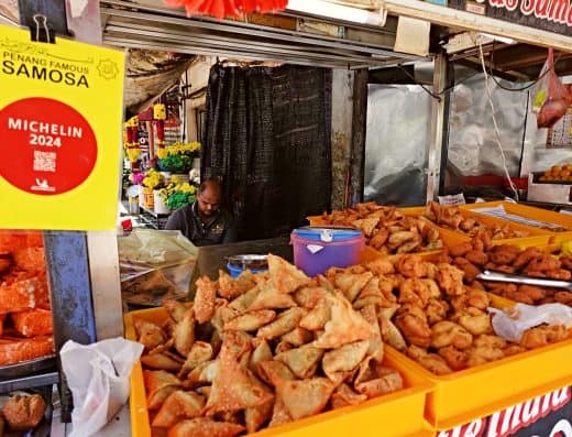 Vibrant street food stall featuring fried snacks, spring rolls, and fried chicken with colorful flowers and signs in the background.