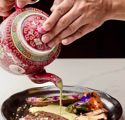 Close-up of a hand pouring a silky sauce onto a beautifully plated gourmet meal with vegetables and edible flowers, showcasing fine dining and culinary artistry at Onalulu.