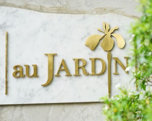 优雅的花园标牌，饰有"au JARDIN"（花园里）字样和蜜蜂图案，采用金色金属材质，安装在郁郁葱葱的户外环境中的大理石墙上。非常适合植物或自然主题空间。.