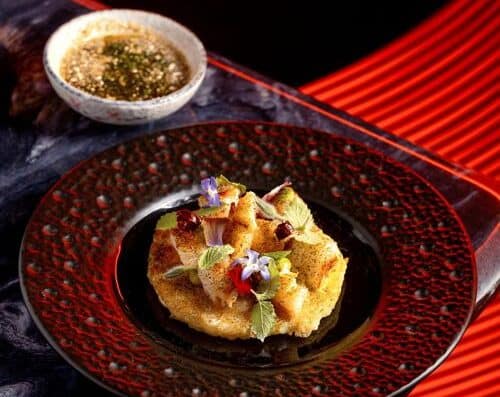 A beautifully plated Japanese tempura appetizer featuring crispy battered vegetables garnished with edible flowers, accompanied by dipping sauce, on a stylish red and black plate.