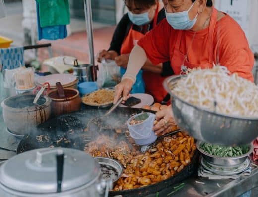 A busy street food stall serving sizzling stir-fried dishes with fresh vegetables, noodles, and savory meats, prepared by skilled cooks wearing face masks, showcasing authentic flavor and vibrant culinary traditions.