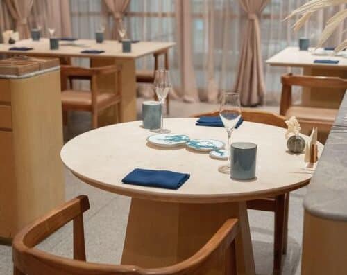 一间温馨明亮、灯光柔和的餐厅里，摆放着时尚的餐桌，蓝色餐巾、香槟酒杯和装饰蜡烛，柔软的窗帘和木制家具营造出温馨的用餐氛围。.